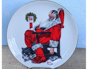 May include: A decorative white plate with a gold rim featuring a painting of Santa Claus resting in a chair. Santa is wearing a red suit with white trim and a red hat. The plate has the text "Dec 26" and the artist's signature.