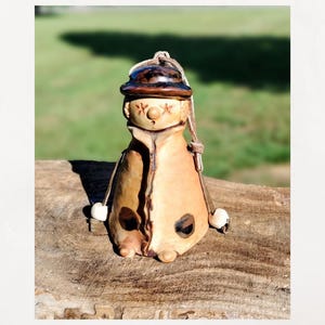 May include: A handcrafted ceramic figurine of a seated figure wearing a hat and coat. The figurine is primarily tan with brown accents, including the hat and a dark spot on the coat. It has a leather cord for hanging and white ball-shaped hands.