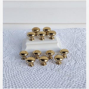 May include: A collection of twelve gold-colored cabinet knobs with a rounded top and a decorative base. The knobs are arranged on a white surface and a white crocheted cloth.