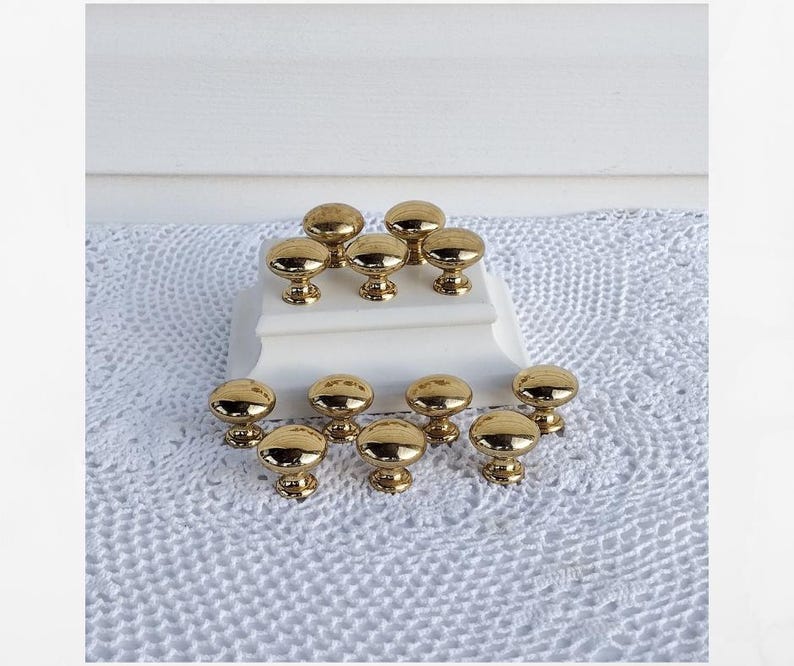 May include: A collection of twelve gold-colored cabinet knobs with a rounded top and a decorative base. The knobs are arranged on a white surface and a white crocheted cloth.