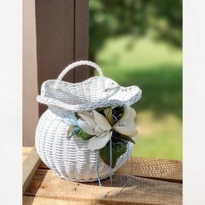May include: A white wicker basket with a handle and a floral arrangement. The basket is round with a ruffled top and is decorated with large white flowers, green leaves, and blue ribbons. The basket is sitting on a wooden surface.