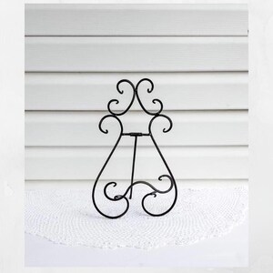 May include: A black metal display stand featuring a decorative scrollwork design. The stand has a central vertical support bar and a curved base with two feet, designed to hold items upright with its symmetrical, open-frame wire construction.