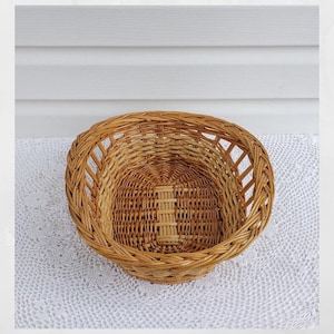 May include: A round, woven wicker basket with a natural brown color. The basket has a wide, open top and a sturdy base. The weave pattern is intricate, with a lattice design around the rim. The basket is empty and sits on a white lace tablecloth.