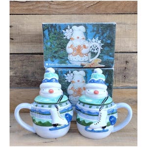 May include: Two ceramic snowman-themed mugs with lids, featuring a blue and green color scheme. Each mug has a snowman head lid and a painted ice skate design. The mugs are in front of a box with a matching design.
