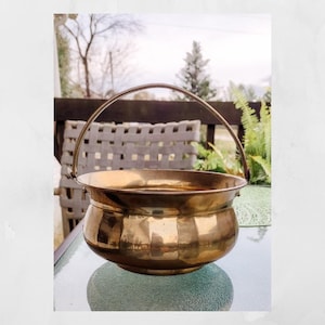 May include: A gold-toned metal planter with a handle. The planter is round and has a slightly flared lip. It is sitting on a glass table.