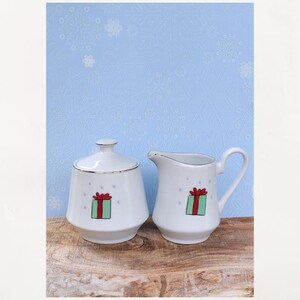 May include: White ceramic sugar bowl with lid and creamer set. Each piece features a green gift box design with a red bow. The rims are trimmed with gold. The background is a light blue with a snowflake pattern.