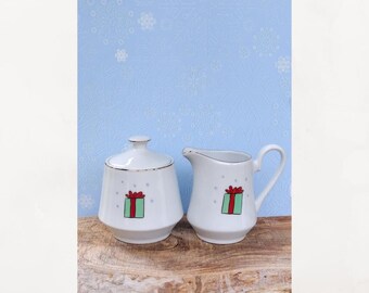 May include: White ceramic sugar bowl with lid and creamer set. Each piece features a green gift box design with a red bow. The rims are trimmed with gold. The background is a light blue with a snowflake pattern.