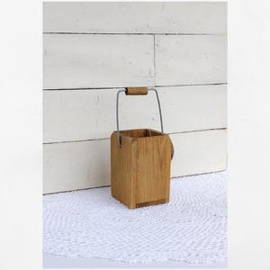 May include: A square wooden bucket with a natural light brown wood grain finish. The container features a thin, silver-colored metal wire handle that is arched upwards, topped with a small, cylindrical wooden grip in the center for carrying.