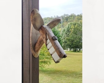 May include: A wall-mounted sculpture crafted from natural wood, featuring a head-like shape with two large, rounded ears, a long snout, and a row of four rectangular white teeth protruding from the lower jaw section.