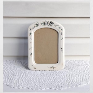 May include: An arched ceramic picture frame with a cream-colored base and a hand-painted botanical motif featuring green leaves, dark berries, and delicate sprigs. The frame has a smooth, glossy finish and a rectangular central opening for a photo.