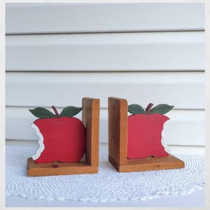 May include: A pair of wooden bookends featuring a red apple design with a bite taken out of the side. Each apple has a dark green leaf on top and is attached to a natural light brown wooden base and vertical support piece for holding books.