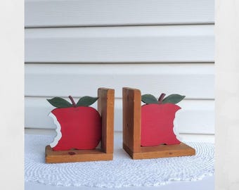 May include: A pair of wooden bookends featuring a red apple design with a bite taken out of the side. Each apple has a dark green leaf on top and is attached to a natural light brown wooden base and vertical support piece for holding books.