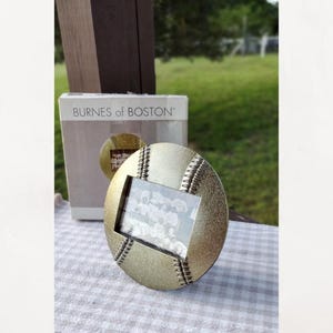 May include: A round, gold-toned metal picture frame shaped like a baseball with textured surface details and raised stitching lines. The frame features a rectangular central opening containing a black and white photograph. A Burnes of Boston box is behind.