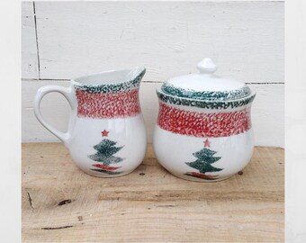 May include: A white ceramic creamer and sugar bowl set. The creamer has a handle and spout, and the sugar bowl has a lid. Both pieces feature a red and green speckled pattern and a Christmas tree design.