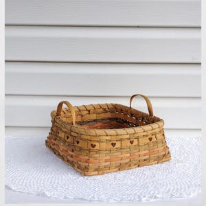 May include: A square woven basket with two arched handles. The basket is made of light brown and peach-colored woven strips. Small heart-shaped cutouts are visible on the front of the basket. The basket is sitting on a white, lacy doily.