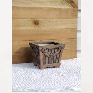 May include: A square-shaped terracotta planter with a dark, weathered finish. The exterior features vertical fluted detailing, decorative floral accents at the corners, and a central leaf-like garland motif. The planter stands on four small feet.