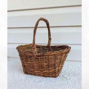 May include: A rectangular brown wicker basket with a woven handle. The basket is made of natural-colored woven material and sits on a white lace surface. The handle is arched and attached to the basket's sides.