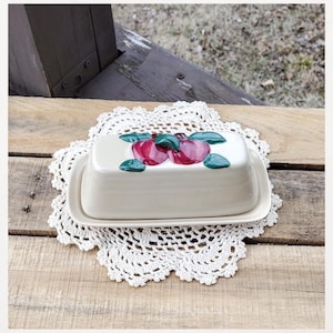 May include: A rectangular, off-white ceramic butter dish with a lid, decorated with a pair of red apples and green leaves. The dish sits on a white, crocheted doily, placed on a wooden surface.