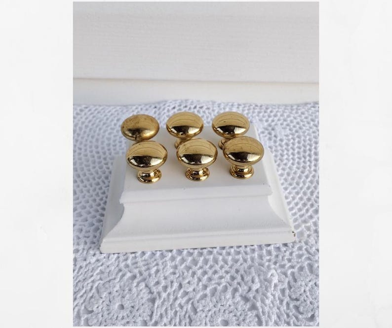 Op de afbeelding: Zes goudkleurige kastknoppen met een afgerond ontwerp worden getoond op een witte, decoratieve basis. De knoppen hebben een glanzende afwerking en zijn gerangschikt in twee rijen van drie. De basis staat op een wit, gehaakt oppervlak.