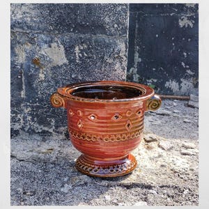 May include: A handcrafted, red-brown ceramic planter with a decorative design. The planter features a wide rim, two scrolled handles, and diamond-shaped cutouts. The base is a pedestal with a detailed, circular pattern.