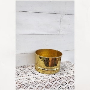 May include: A hammered brass planter with a rounded shape. The planter has a shiny gold finish and a textured surface. It sits on a white lace doily in front of a white wooden background.