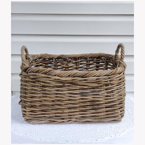 May include: A rectangular woven basket made of natural brown willow branches. The basket features a thick, braided rim and two sturdy, integrated handles on the left and right sides. The weave pattern is open and rustic, showing varied brown tones.