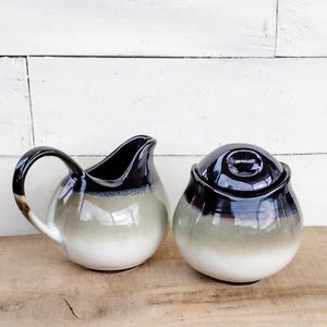 May include: A ceramic creamer and sugar bowl set. The creamer has a curved handle and spout, while the sugar bowl has a lid. Both pieces are glazed in a gradient of colors, transitioning from white to gray and black.
