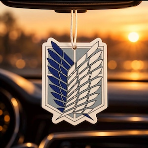 Puede incluir: Un ambientador de coche colgante con el emblema de las Alas de la Libertad. El diseño en forma de escudo muestra un gráfico dividido con un ala azul oscuro a la izquierda y un ala blanca a la derecha, sobre un fondo gris claro con bordes negros.