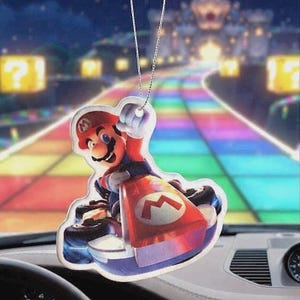 May include: A hanging car air freshener featuring a colourful graphic of Mario driving a red go-kart with a white M logo on the front. The character wears a red cap and blue overalls, with one hand raised. It hangs from a thin silver chain.