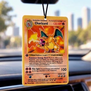 May include: A rectangular car air freshener featuring a Charizard Pokémon trading card design. The card displays the orange dragon-like creature breathing fire, with 120 HP, the text Charizard, and game statistics printed in black on a yellow background.