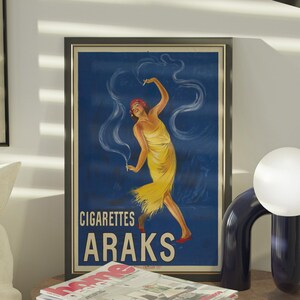 May include: A room interior with framed vintage posters and a modern lamp. One poster advertises "Cigarettes Araks" with a dancing woman in yellow. A stack of magazines sits on a round, textured side table. A white globe lamp and a black floor lamp are also visible.