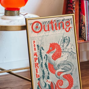 May include: A framed vintage illustration of a seahorse with a man riding it. The illustration is in red, blue, and green colours. The text "Outing for April" is printed in red on the poster.