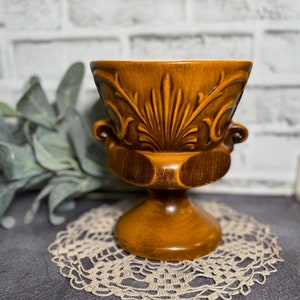 May include: A brown ceramic planter with a decorative, ornate design. The planter has a wide, round base and a smaller, round top. The planter is sitting on a white doily.