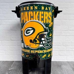 以下が含まれることがあります： 黒いベースと蓋が付いたグリーンベイパッカーズのコーヒーurn。 チームのロゴ、緑色の背景に緑色の「G」が付いた黄色のヘルメットで装飾されています。 テキスト「GREEN BAY PACKERS」は黄色で表示されます。
