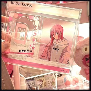 May include: A clear acrylic display stand featuring the Blue Lock character Hyoma with pink hair, wearing a light-colored hoodie. The stand includes a rectangular base with the name HYOMA printed in black, alongside soccer ball graphics.