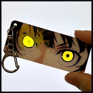 May include: A rectangular acrylic keychain featuring a close-up illustration of a pair of intense, glowing yellow eyes with dark pupils set against a dark-haired character's face. The keychain includes a silver-toned metal lobster clasp attachment.