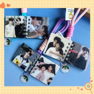May include: Small photo albums with spiral binding, featuring images of people. Each album is attached to a lanyard with a pink and purple cord. The background is a light blue surface with a yellow border.