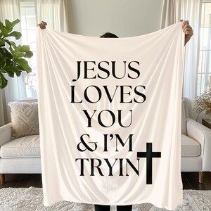 Puede incluir: Manta color crema con el texto negro "JESUS LOVES YOU & I'M TRYIN" y una cruz negra. La manta se sostiene, mostrando el mensaje.