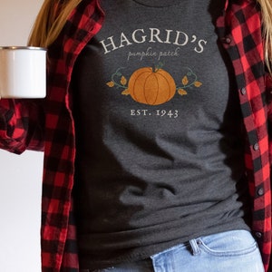 May include: Dark gray t-shirt with the text "Hagrid's Pumpkin Patch" and a pumpkin graphic. The shirt is paired with a red and black plaid flannel shirt and blue jeans. A white mug is held in the hand.
