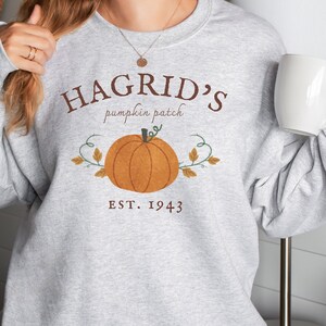 Könnte beinhalten: Ein graues Sweatshirt mit einem braunen Grafikdesign, auf dem "Hagrid's pumpkin patch est. 1943" mit einem Cartoon-Kürbis und Ranken steht.