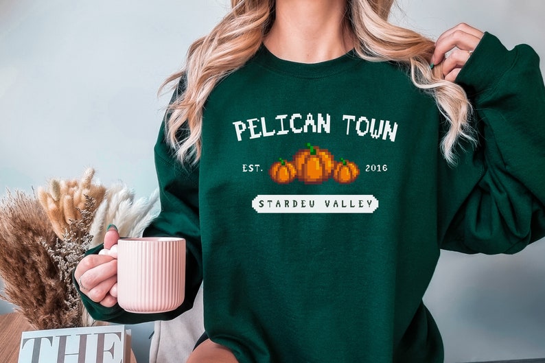 May include: Green sweatshirt with a white graphic that says "Pelican Town Est. 2016 Stardew Valley" with a pixelated image of pumpkins.