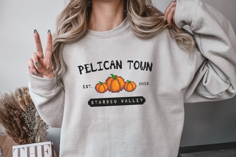May include: A gray sweatshirt with a black graphic print that says "Pelican Town Est. 2016 Stardew Valley" with a pixelated image of three pumpkins.