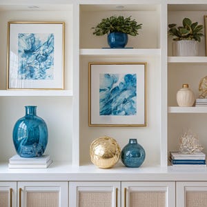 Könnte beinhalten: Ein weißer Einbauschrank mit offenen Regalen, der dekorative Gegenstände präsentiert. Die Regale zeigen gerahmte blaue abstrakte Kunst, teal Vasen, goldene Akzente und Topfpflanzen. Die Schranktüren haben gewebte Paneele und goldene Griffe.