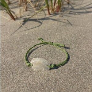 May include: A bracelet featuring a single, smooth, off-white seashell centrepiece threaded onto a thin, olive green cord. The cord includes adjustable sliding knots on each side of the shell, creating a simple, circular band design.