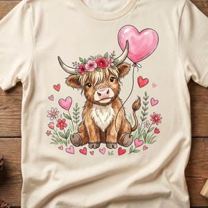 Può includere: T-shirt color crema con un cartone animato di mucca delle Highlands con una corona floreale, che tiene in mano un palloncino rosa a forma di cuore. Il design include cuori rosa, fiori e foglie verdi. La mucca è marrone e bianca.