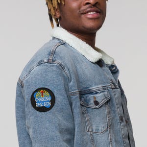May include: Light blue denim jacket with a white sherpa collar. The jacket features a round patch on the left sleeve with the text "CHASING THE SON" in blue and yellow. The jacket has two chest pockets and a button closure.