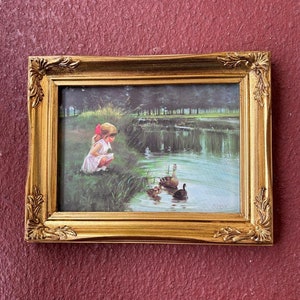 May include: A framed painting of a young girl in a white dress and red bow feeding ducks in a pond. The painting is set in a green meadow with trees in the background. The frame is gold with ornate details.