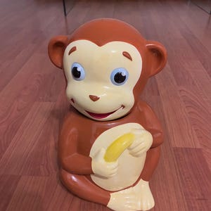 May include: A ceramic monkey figurine with a brown body, tan face, and blue eyes. The monkey is holding a yellow banana. The figurine is sitting on a wooden floor.