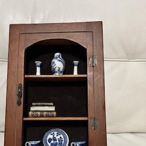 May include: A wooden display cabinet with a glass door and two shelves. The cabinet is filled with miniature porcelain items, including a vase, cups, and a plate, all decorated with a blue and white floral pattern. There are also a few small books on the lower shelf.