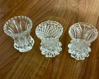 May include: Three small, clear glass bud vases with a diamond-shaped pattern. The vases are on a wooden surface.
